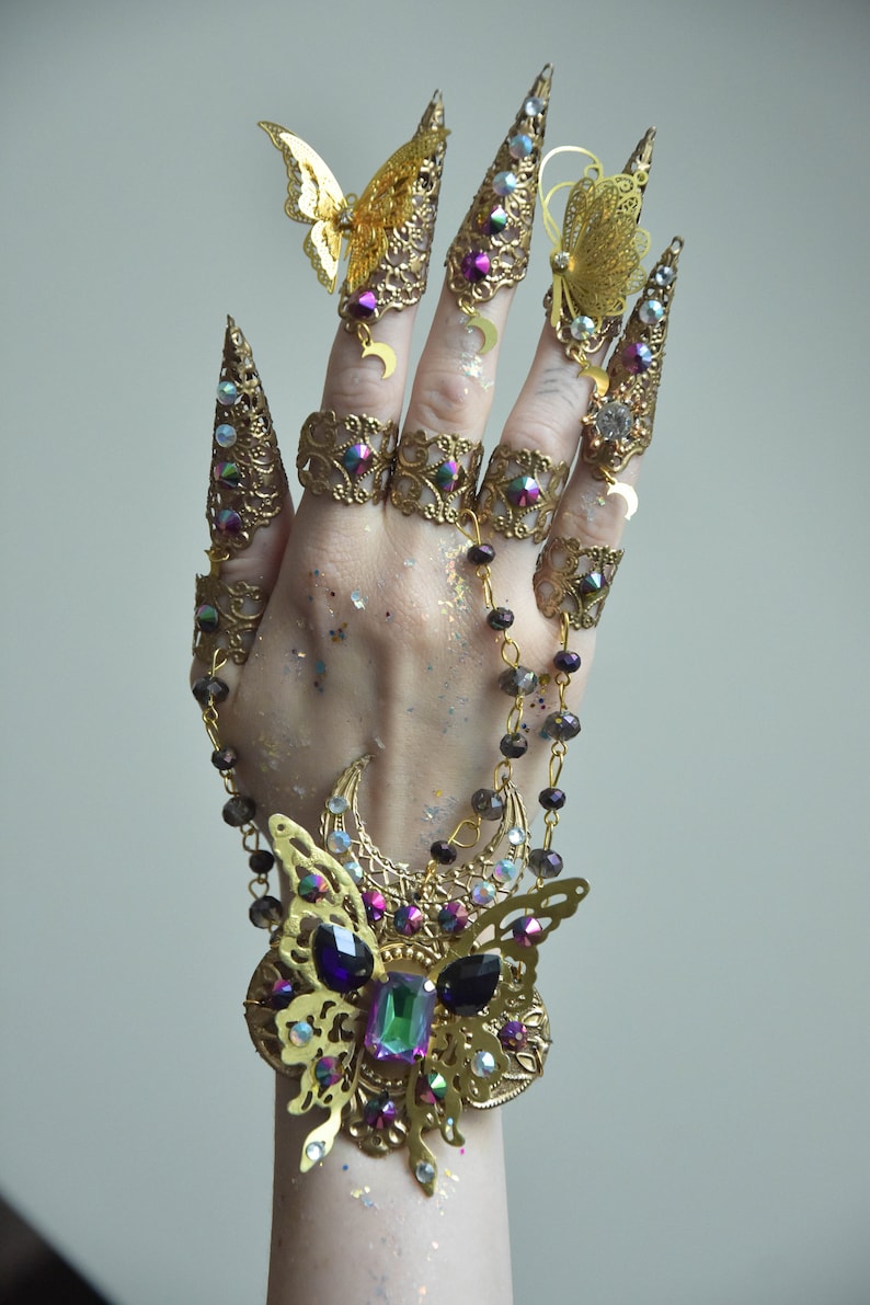 May include: A gold-toned finger ring set with a butterfly design and iridescent gemstones. The set includes multiple rings with a crescent moon design and a chain with iridescent beads.
