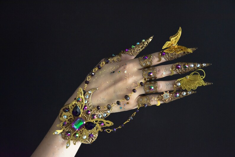 May include: A gold-toned butterfly-themed finger and wrist jewelry set. The set includes a bracelet with a large butterfly design and several rings with butterfly and claw designs. The jewelry is adorned with colorful gemstones.