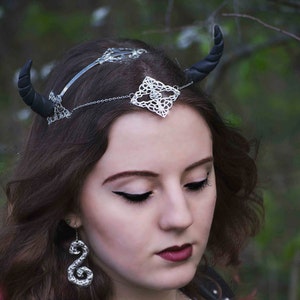 May include: A woman with dark brown hair is wearing a silver headband with black horns and a silver filigree design. She is also wearing silver earrings with a spiral design.