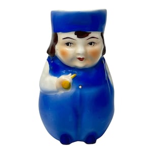 Może przedstawiać: Figurka ceramiczna w stylu vintage w kształcie osoby w niebieskim mundurze i czapce. Figurka ma malowaną twarz z różowymi policzkami, brązowymi włosami i czerwonymi ustami. Trzyma żółty przedmiot.