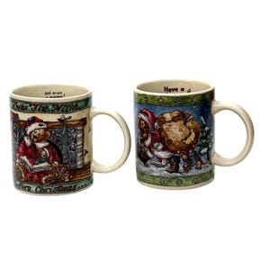 May include: Two ceramic Christmas mugs with cream-colored handles. One mug features a bear in a Santa suit reading a book by a fireplace. The other mug depicts a bear carrying a sack of gifts in a snowy scene. Both mugs have festive text.