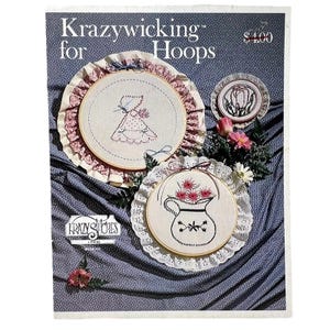 May include: A vintage craft pattern titled "Krazywicking for Hoops" featuring embroidered designs. The image showcases three hoop art examples: a dress, a floral pitcher, and a tulip design. The patterns are surrounded by decorative lace and floral accents, set against a blue patterned background.