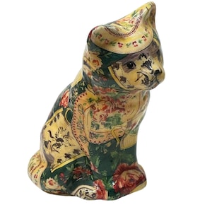 May include: Ceramic cat figurine with a floral pattern in shades of green, yellow, and red. The cat is sitting upright with its head turned slightly to the side. The design features roses and other flowers.