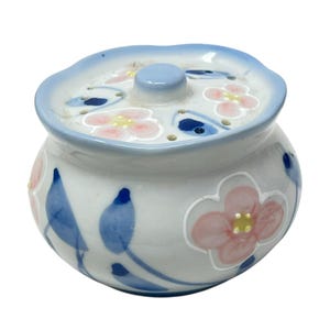 Może przedstawiać: Mały, okrągły ceramiczny pojemnik z pokrywką. Pojemnik jest biały z niebieskimi i różowymi kwiatowymi wzorami. Obręcz i pokrywa są jasnoniebieskie. Pokrywa ma mały, okrągły guziczek pośrodku.