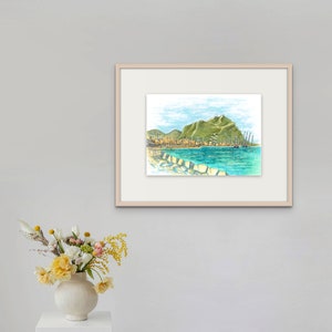 May include: A framed watercolor painting of a coastal town with a green hill in the background. The town is on the shore of a blue body of water with boats in the water. The painting is in a light wood frame.