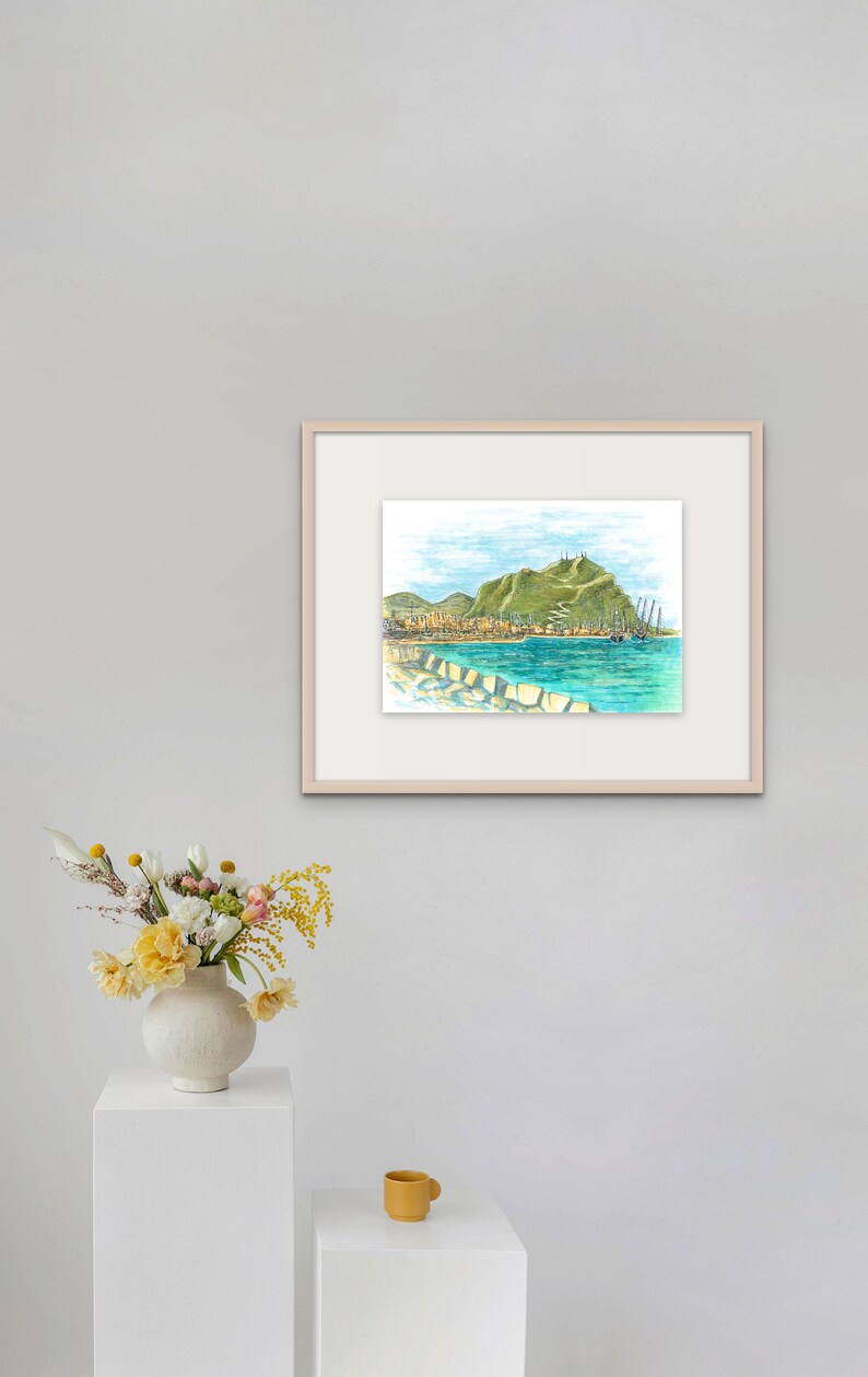 May include: A framed watercolor painting of a coastal town with a green hill in the background. The town is on the shore of a blue body of water with boats in the water. The painting is in a light wood frame.