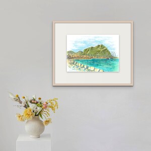 May include: A framed watercolor painting of a coastal town with a green hill in the background. The town is on the water and there are boats in the water. The painting is in a light wood frame.