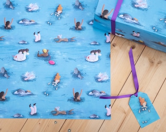 Könnte beinhalten: Blaues Geschenkpapier mit einem sich wiederholenden Muster von Menschen, die im Meer schwimmen. Das Papier ist um eine Geschenkbox mit einem lila Band und einem Geschenkanhänger mit einer Meerjungfrauen-Illustration gewickelt.