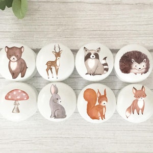 May include: Eight white ceramic knobs with woodland animal and mushroom designs. The knobs feature a bear, deer, raccoon, hedgehog, mushroom, rabbit, squirrel, and fox.