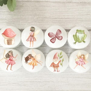 May include: Set of eight white ceramic knobs with whimsical woodland creature and fairy designs. The designs include a mushroom house, a butterfly, a frog, a gnome, and fairies.