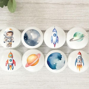 May include: Eight round white ceramic knobs with space-themed illustrations. The designs include an astronaut, a rocket ship, a planet, and a starry sky.