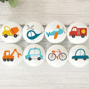 May include: Eight white round knobs with colourful cartoon images of vehicles, including a cement truck, helicopter, aeroplane, fire engine, excavator, police car, bicycle, and car.