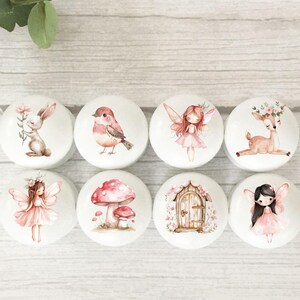 May include: Eight round white ceramic knobs with various woodland creature and fairy designs. The designs include a rabbit, bird, fairy, deer, mushroom, door, and two fairies.