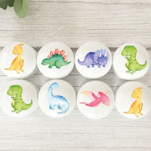 May include: Eight white ceramic knobs featuring different dinosaur illustrations. The dinosaurs are painted in various colours, including green, yellow, blue, purple, and pink.