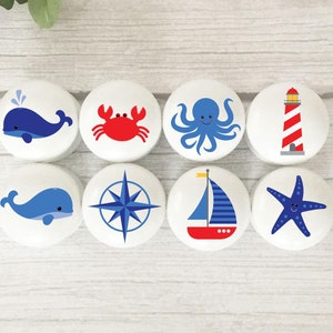 May include: Set of eight nautical themed knobs with various designs including a whale, crab, octopus, compass, sailboat, starfish, and lighthouse. The knobs are white with blue, red, and yellow accents.