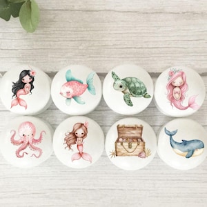 May include: Eight round white ceramic knobs with various watercolour illustrations of sea creatures and mermaids. The illustrations include a mermaid with pink hair, a mermaid with brown hair, a pink fish, a green sea turtle, a pink octopus, a treasure chest, a blue whale, and a mermaid with a crown of flowers.