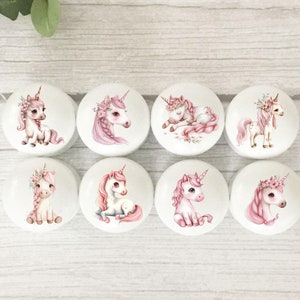 May include: Eight white round knobs with pink unicorn illustrations. Each knob features a different unicorn pose with pink floral accents.