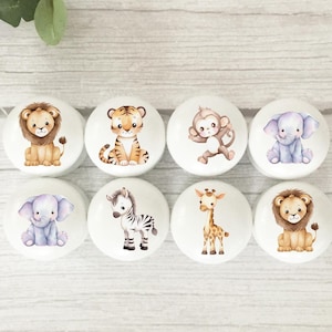 May include: Eight white cabinet knobs featuring watercolour illustrations of safari animals, such as a lion, tiger, monkey, elephant, zebra, and giraffe. The knobs are arranged in two rows of four, suitable for a nursery or children's room.