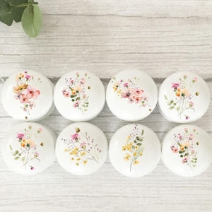 May include: Eight white ceramic knobs with floral designs. The knobs feature watercolour-style flowers in shades of pink, yellow, and green.