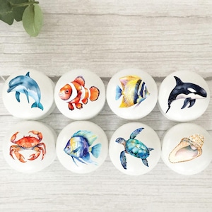 May include: Set of eight white ceramic knobs with colourful ocean-themed illustrations. The designs include a dolphin, clownfish, angelfish, orca, crab, blue tang, sea turtle, and seashell.