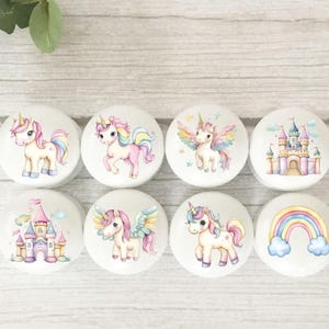 May include: Eight white cabinet knobs with unicorn and castle designs. Each knob features a different illustration with pastel colours, including pink, yellow, and blue. The knobs are round and have a smooth finish.