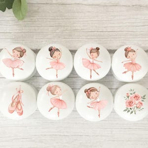 May include: Eight white ceramic cabinet knobs with ballerina designs. Each knob features a different ballerina pose or a pair of ballet shoes. The ballerinas wear pink tutus and have brown hair. One knob has a bouquet of pink roses.