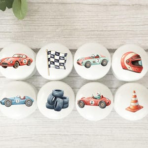 May include: Eight white ceramic knobs with racing car designs. The illustrations include race cars, a chequered flag, tyres, a helmet, and a traffic cone. The knobs feature red, blue, and green accents, suitable for a child's room or a racing fan.