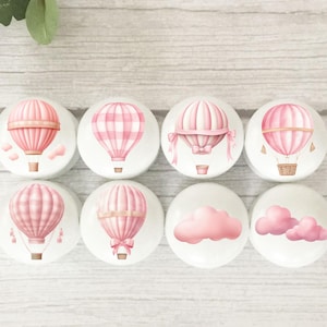 May include: Eight white cabinet knobs with pink hot air balloon and cloud designs. The balloons feature various patterns and details, including stripes, bows, and baskets. The knobs are arranged on a light wood surface.