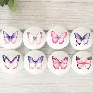 May include: Eight white, round cabinet knobs with butterfly designs. Each knob features a different butterfly with various colours, including pink, purple, and blue. The knobs are arranged in two rows of four.