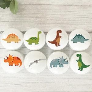 May include: Set of eight white ceramic drawer knobs featuring colourful dinosaur illustrations. The dinosaurs include a stegosaurus, triceratops, pterodactyl, brachiosaurus, and a tyrannosaurus rex.