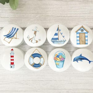 May include: Eight white ceramic cabinet knobs with nautical designs. Each knob features a different watercolour illustration: a beach chair, anchor, sailboat, beach hut, lighthouse, life preserver, bucket of shells, and a dolphin.