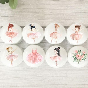 May include: Eight round white ceramic knobs with various designs featuring pink ballerina figures, a pink tutu, and a floral bouquet.