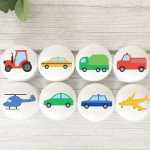 May include: Eight white cabinet knobs with colourful vehicle designs. The knobs feature a red tractor, yellow taxi, green truck, red fire truck, blue helicopter, green car, blue car, and yellow aeroplane.