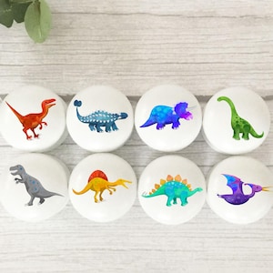 May include: Eight white cabinet knobs, each decorated with a different dinosaur illustration. The dinosaurs are depicted in various colours, including red, blue, green, yellow, and purple. The knobs are arranged in two rows of four.