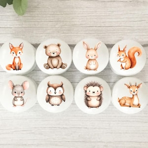 May include: Set of eight round wooden knobs featuring cute animal illustrations. The animals include a fox, bear, rabbit, squirrel, mouse, owl, hedgehog, and deer. The knobs are painted in shades of brown, orange, and white.