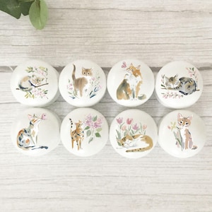 May include: A set of eight white ceramic cabinet knobs, each adorned with a hand-painted cat design. The knobs showcase different cat illustrations, complemented by floral motifs. The cats are in various poses and colours, offering a charming and whimsical appearance.