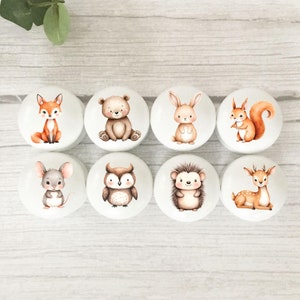 May include: A set of eight round white cabinet knobs, each featuring a different watercolour-style illustration of a woodland animal, including a fox, bear, rabbit, squirrel, mouse, owl, hedgehog, and deer, arranged in two rows of four.