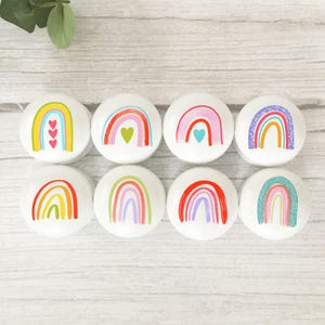 May include: Eight white cabinet knobs, each decorated with a different rainbow design. The rainbows are in various colours, including yellow, red, pink, blue, and green, with heart details. The knobs are displayed on a light wooden surface.