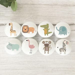 May include: Eight white cabinet knobs with animal illustrations. The knobs feature a giraffe, lion, crocodile, elephant, hippo, flamingo, monkey, and zebra. The knobs are round and have a glossy finish.