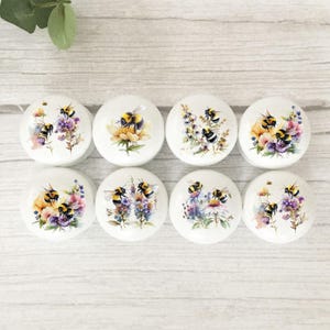 May include: Eight white ceramic cabinet knobs with bee and floral designs. Each knob features a different arrangement of bumblebees and colourful flowers, including yellow, purple, and pink blooms. The knobs are round and have a glossy finish.