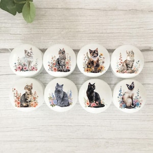May include: Eight white ceramic cabinet knobs featuring watercolour cat illustrations surrounded by flowers. The cats are in various colours and patterns, including grey, black, and tortoiseshell. Knobs are on a light wood surface.