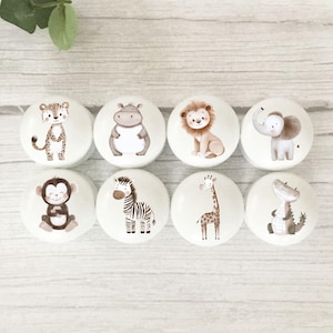 May include: A set of eight white ceramic cabinet knobs, each decorated with a different safari animal illustration. The collection includes a leopard, hippo, lion, elephant, monkey, zebra, giraffe, and crocodile. The knobs are round and glossy.