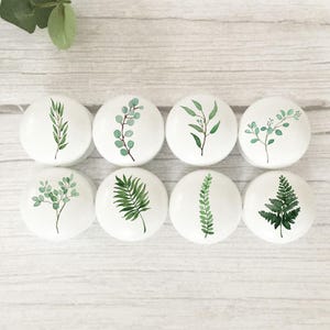 May include: Set of eight white ceramic knobs with green watercolour leaf designs. The knobs feature various leaf shapes, including ferns, eucalyptus, and palm leaves.