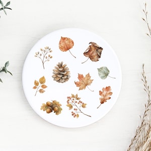 May include: A round white ceramic coaster featuring a watercolour illustration of various autumn elements including a pinecone, acorns, berries, and assorted brown, orange, and green leaves arranged in a scattered pattern across the surface.