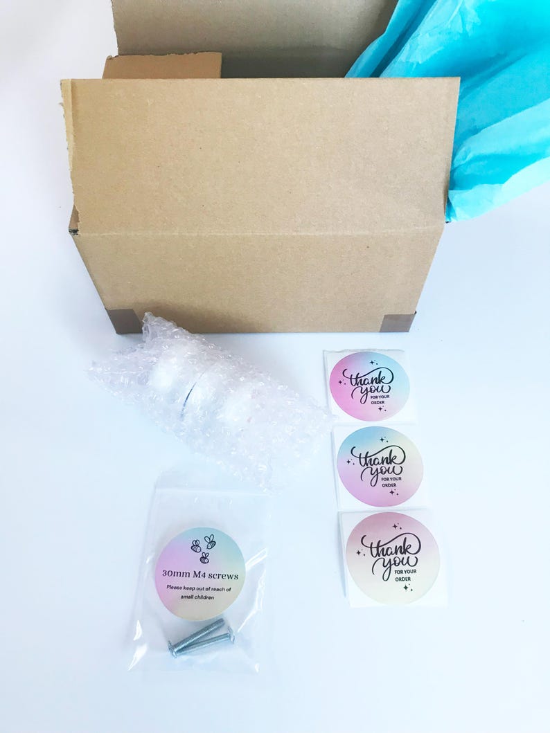 Op de afbeelding: Een kartonnen doos met blauwe tissuepapier vulling. De doos bevat een witte knop, drie zilveren schroeven en drie bedank stickers met een regenboogkleurige achtergrond. De stickers zeggen "Bedankt voor uw bestelling".