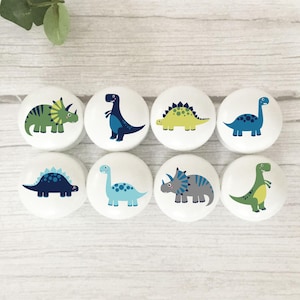 May include: Eight white ceramic cabinet knobs with colourful dinosaur illustrations. Each knob features a different dinosaur design in shades of green, blue, and yellow. The knobs are arranged on a light wood surface.