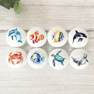 May include: Eight white ceramic cabinet knobs with marine life illustrations. Designs include a blue dolphin, clownfish, yellow and blue fish, orca, red crab, blue fish, sea turtle, and a grey seal. Ideal for nautical or ocean-themed decor.