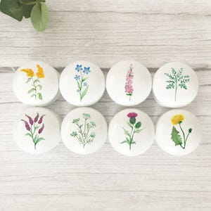 May include: Eight white ceramic cabinet knobs, each featuring a hand-painted floral design. The flowers are in shades of yellow, blue, pink, and purple, with green leaves. The knobs are displayed on a light wood-effect surface.