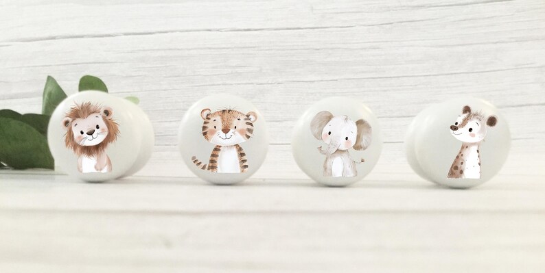 以下が含まれることがあります： 動物のイラストが描かれた白いセラミック製の引き出しノブが4つ。ノブには、ライオン、トラ、ゾウ、キリンが描かれています。