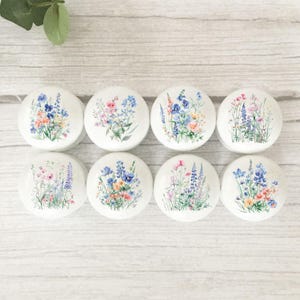 May include: Eight white cabinet knobs with floral designs. Each knob features a different arrangement of colourful flowers, including blue, pink, and yellow blooms, set against a white background. The knobs are arranged on a light wood surface.